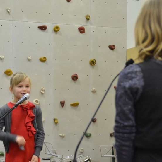 Ein kleines Mädchen in einem roten Kleid spricht in ein Mikrofon vor einer Kletterwand.