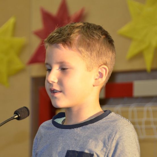 Ein junger Junge steht vor einem Mikrofon, wahrscheinlich bei einer Schulveranstaltung. Er trägt ein graues Shirt und hat kurze blonde Haare. Hinter ihm zieren dekorative Papiersterne und Blätter die Wand.