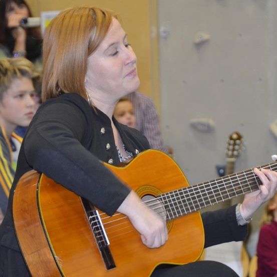 Eine Frau mit braunen Haaren spielt Gitarre, während sie in einem Raum sitzt und Kinder zusehen. Ein Kind scheint zu schlafen.