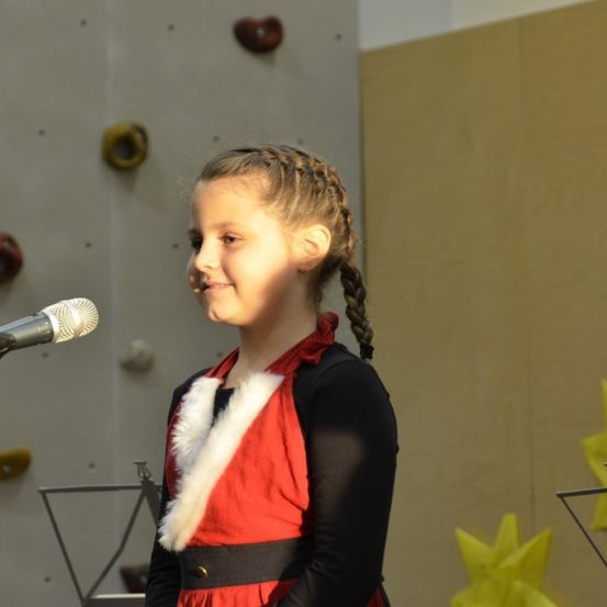 Ein junges Mädchen steht vor einem Mikrofon, gekleidet in einem roten und schwarzen Kostüm mit weißem Pelzbesatz. Hinter ihr ist eine Kletterwand mit verschiedenen Formen und Farben zu sehen.