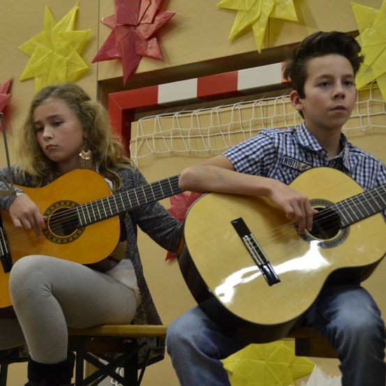 Zwei Kinder sitzen auf einer Bank und spielen Gitarre. Ein Mädchen links und ein Junge rechts, beide konzentrieren sich auf ihre Instrumente. Sie befinden sich in einem Raum, der mit Papiersternen dekoriert ist.