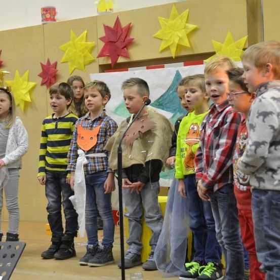 Kinder stehen in einer Gruppe und singen, während eines in ein Mikrofon spricht. Einige von ihnen tragen Brillen, Turnschuhe und Stiefel. Ein Banner hängt hinter ihnen an der Wand.