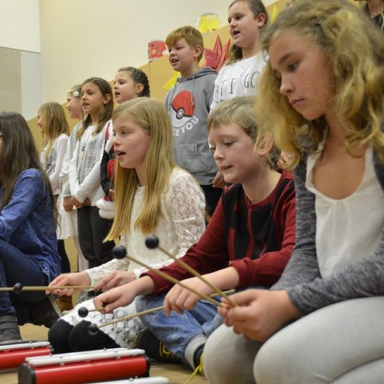 Eine Gruppe von Kindern sitzt auf dem Boden in einem Klassenzimmer. Sie spielen Xylophon. Das Mädchen vorn trägt eine Brille und Turnschuhe. Der Junge neben ihr trägt ein rotes Shirt. Hinter ihnen stehen andere Kinder. Eines von ihnen trägt eine graue Kapuzenjacke.