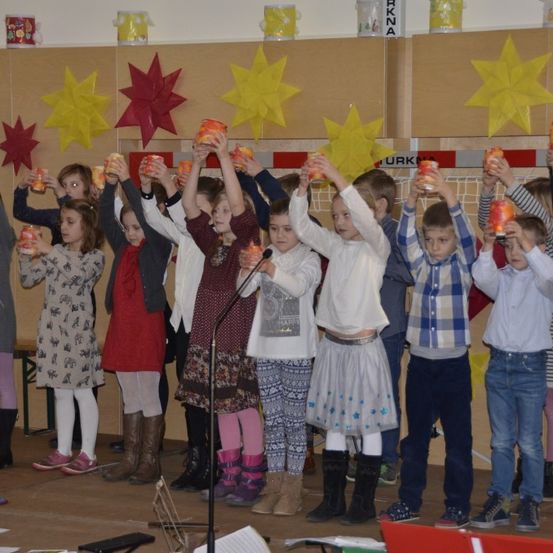 Eine Gruppe von Kindern auf der Bühne hält Tassen in den Händen und nimmt wahrscheinlich an einer festlichen Veranstaltung teil, an der gelbe Sterne an der Wand hängen.