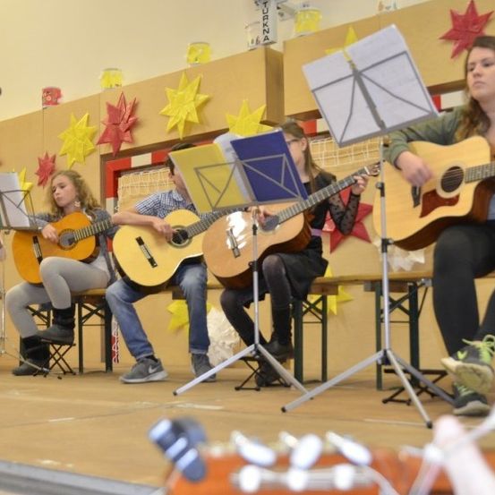 Eine Gruppe junger Musiker tritt mit ihren Gitarren auf der Bühne auf, begleitet von Notenständern. Sie sitzen auf Klappstühlen, mit dekorativen Sternen an der Wand dahinter.