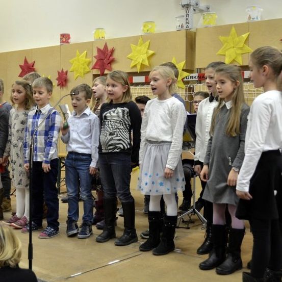 Eine Gruppe von Kindern, in Winterkleidung gekleidet, tritt auf der Bühne auf. Sie halten Mikrofone und singen gemeinsam. Hinter ihnen befindet sich eine dekorierte Kulisse mit Sternen und einem Holzregal mit verschiedenen Gegenständen.