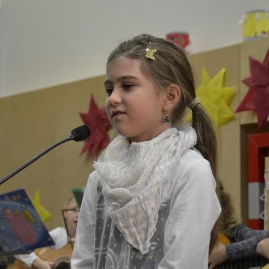 Ein junges Mädchen steht vor einem Mikrofon und singt während einer Aufführung mit Gitarren im Hintergrund. Der Hintergrund zeigt eine Wand mit roten Sternendekorationen.