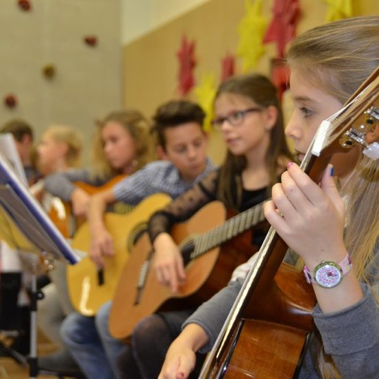 Eine Gruppe von Kindern spielt Gitarren in einem Klassenzimmer. Ein Mädchen vorne hat eine pinke Uhr am Handgelenk.