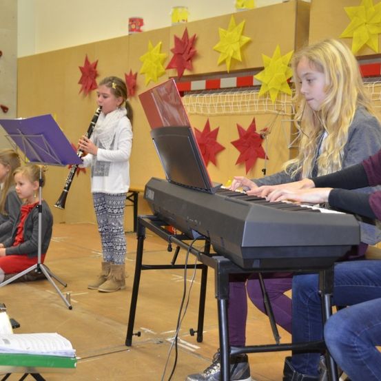Mehrere Kinder in einem Raum mit dekorativen Sternen an der Wand spielen Instrumente. Ein Mädchen spielt Klarinette, während ein anderes das Klavier spielt.