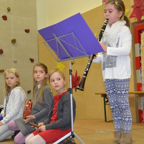 Ein Mädchen spielt eine Klarinette in einem Klassenzimmer, wobei drei andere Mädchen hinter ihr sitzen und Instrumente halten.