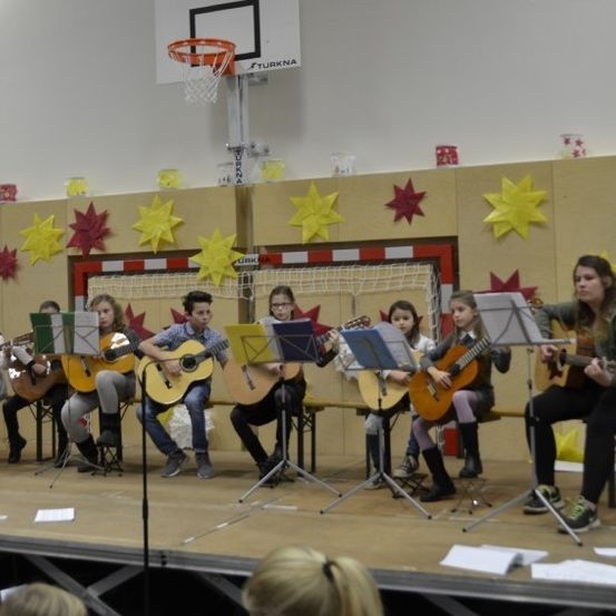 Eine Gruppe von Kindern spielt auf der Bühne mit Gitarren. Einige sitzen, andere stehen. Sie sind in einem Raum, der mit Sternen und einem Basketballkorb dekoriert ist.