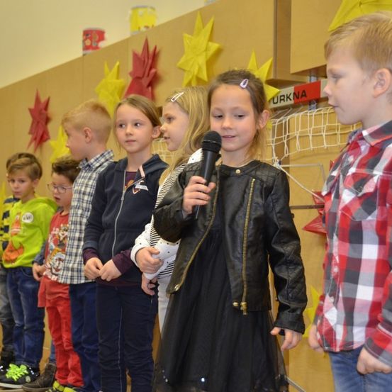 Eine Gruppe von Kindern, darunter ein Mädchen mit einem Mikrofon, steht in einer Reihe auf einer Bühne mit einem braunen Hintergrund, der mit roten Sternen verziert ist.