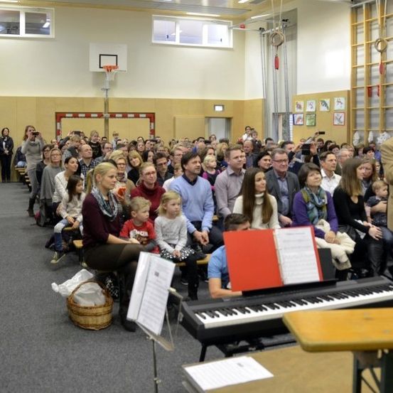 Ein Mann dirigiert einen Chor vor einem Keyboard, während eine Menschenmenge in einem großen Raum hinter ihnen sitzt.