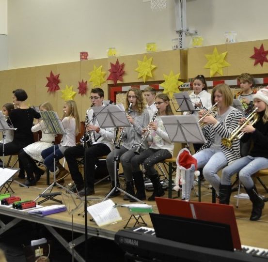 Eine Gruppe von Kindern und Jugendlichen spielt ein Musikstück mit Instrumenten auf einer Bühne. Sie sitzen auf Stühlen mit Notenständern und spielen Trompeten, Flöten und andere Instrumente.