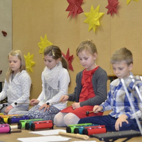 Vier Kinder sitzen auf dem Boden und spielen Xylophon. Hinter ihnen eine mit sternförmigen Dekorationen verzierte Wand.