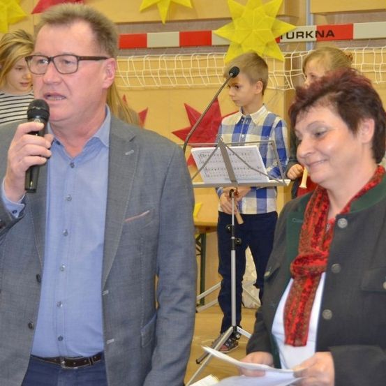 Ein erwachsener Mann hält ein Mikrofon beim Sprechen und steht neben einer Frau, die Papiere in den Händen hält. Dahinter versammeln sich Kinder um einen Notenständer mit einem Buch. Ein Junge steht neben dem Ständer, trägt eine Brille. Im Hintergrund ist eine Wand mit sternförmigen Papieren verziert.