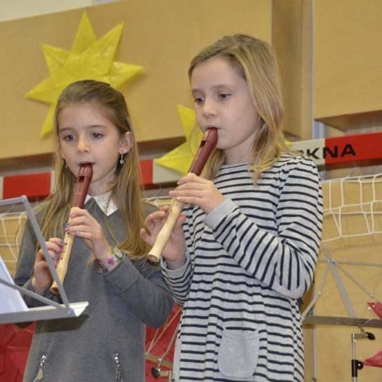 Zwei junge Mädchen spielen Holzflöten in einem Raum mit gelben Sternen und einem roten Banner, das 'KNA' steht. Ein Mädchen trägt ein graues Sweatshirt, und das andere trägt ein gestreiftes Kleid.