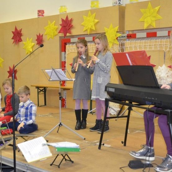 Eine Gruppe von Kindern tritt auf der Bühne auf, zwei Mädchen spielen Flöten, ein Mädchen spielt Keyboard und andere sitzen mit Musikinstrumenten. Hinter ihnen ist eine Wand mit Papiersternen und einem Netz.