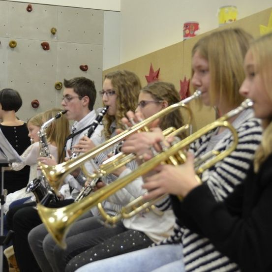 Eine Gruppe junger Musiker übt in einem Raum mit Musikinstrumenten. Die Instrumente umfassen Trompeten, eine Saxophon und eine Flöte. Sie tragen Brillen und eine Frau im Hintergrund liest Notenpapiere.