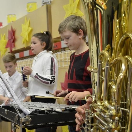 Mehrere Kinder üben Musik. Ein Junge spielt die Tuba, während ein Mädchen das Xylophon spielt. Ein weiterer Junge hält eine Gitarre. Dahinter sind einige Papiersterne an der Wand befestigt.