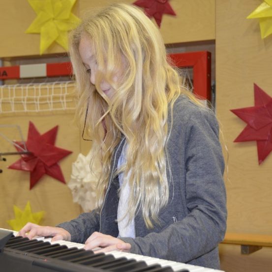 Ein junges Mädchen mit blonden Haaren spielt Klavier. Sie trägt eine graue Jacke. Hinter ihr befinden sich rote Sternendekorationen und ein gelbes Banner mit dem Wort 'KNA'. Ein roter Rahmen ist an der Wand befestigt.