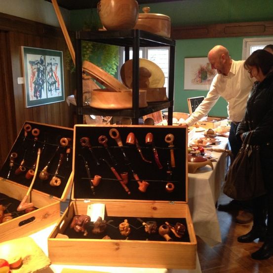 Eine Indoor-Ausstellung von Holzgegenständen, einschließlich Schüsseln, Tellern und Pfeifen, auf Regalen und Tischen angeordnet. Ein Mann und eine Frau betrachten die Gegenstände. Der Raum hat Holzwände und Gemälde.