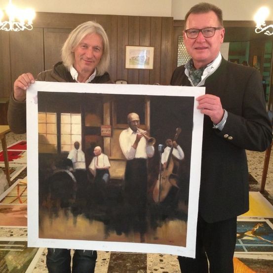 Zwei Männer halten ein großes gerahmtes Gemälde, das eine Band darstellt. Die Band besteht aus Musikern, die Trompete, Saxophon und Cello spielen. Sie befinden sich in einem Raum mit Holzwänden und einem Kronleuchter.