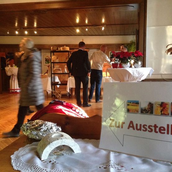 Ein Raum mit Holzböden, zwei Personen in Anzügen, eine Frau in einem Mantel und ein Schild, das Zur Austellung sagt. Es gibt Tische, Blumenvasen und ausgestellte Gegenstände.