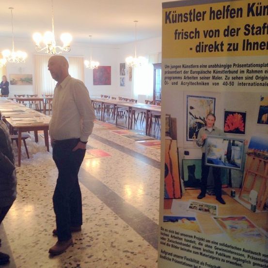 Eine Kunstausstellung mit einem Mann, der einen Rahmen hält, vor einem Banner stehend. Das Banner hat deutschen Text. Eine Person steht links, in der Nähe von Tischen mit Papieren. Kronleuchter hängen von der Decke.