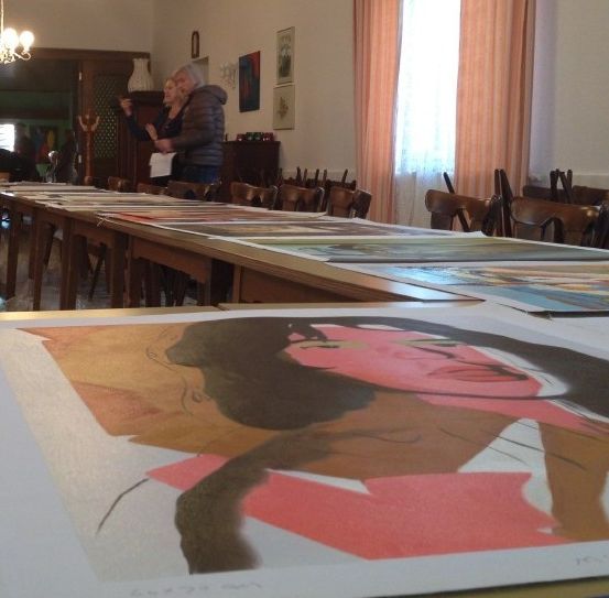 Ein Raum mit mehreren Gemälden auf Tischen, einer Deckenleuchte und zwei Personen, die an einer Wand mit gerahmten Bildern und einer Vase stehen.