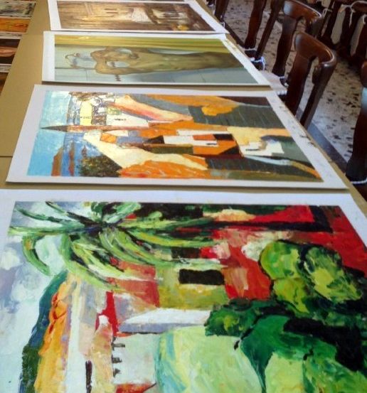 Ein Tisch mit mehreren Gemälden, darunter abstrakte und realistische Stile, ist in einer Reihe angeordnet. Stühle sind um den Tisch herum aufgestellt.