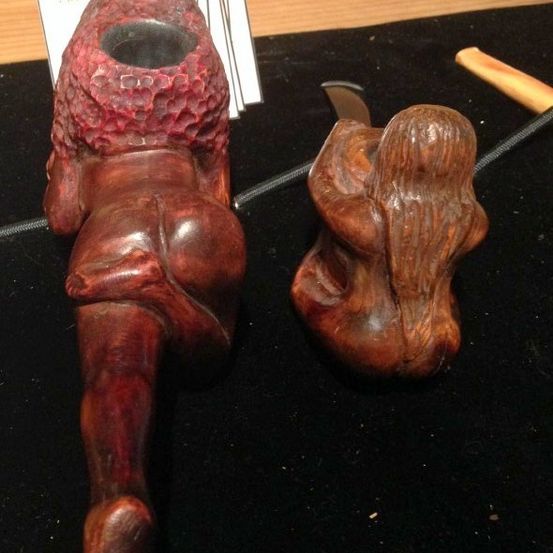 Zwei hölzerne Skulpturen auf einer schwarzen Samtfläche, eine größere, die eine Figur in kauernder Position darstellt, und eine kleinere sitzende Figur. Eine Pfeife und ein Messer sind in der Nähe.