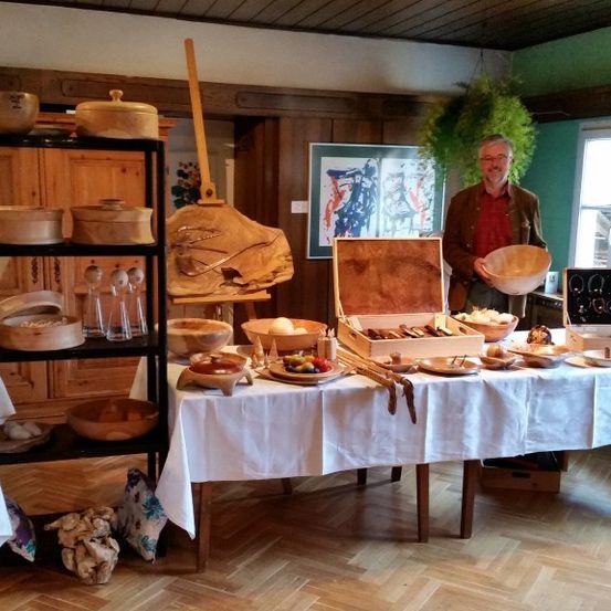 Ein Mann steht hinter einem Tisch mit Holzschüsseln und Utensilien. Verschiedene Holzgegenstände befinden sich auf Regalen und in Kisten. Ein Gemälde ist an der Wand.