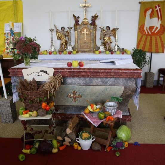 Ein Altar mit verschiedenen Früchten, Gemüse und religiösen Symbolen, umgeben von einem roten Teppich und einem Holzstuhl.