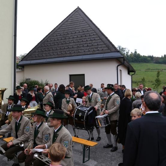 Eine Gruppe von Musikern in grünen Uniformen spielt Instrumente vor einer Menschenmenge in der Nähe eines Hauses mit einem dreieckigen Dach.