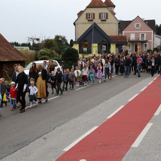 Eine große Gruppe von Menschen, darunter Kinder in Kostümen, geht unter einem bewölkten Himmel eine Straße entlang, mit Gebäuden auf beiden Seiten.