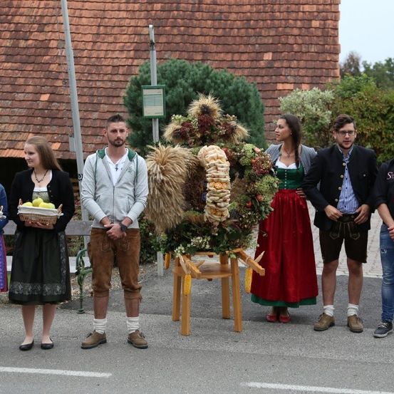 Eine Gruppe von Menschen, gekleidet in traditioneller deutscher Tracht, steht um einen geschmückten Wagen auf einem Stadtplatz. Einige halten Körbe mit Obst, während andere in Gespräche vertieft sind.