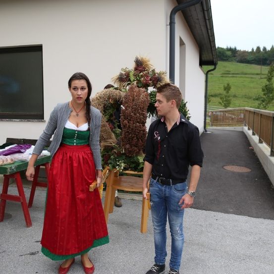 Eine Frau in einem traditionellen roten und grünen Kleid steht neben einem Mann in lässiger Kleidung. Sie stehen vor einem Haus mit Garten.