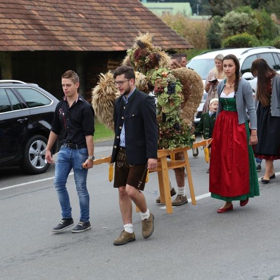 Menschen gehen die Straße entlang, darunter zwei Männer und zwei Frauen in traditioneller bayerischer Tracht. Eine Löwenstatue wird auf einer Holzplattform getragen.