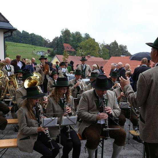 Eine Musikergruppe in traditioneller Kleidung spielt im Freien Instrumente, mit Zuschauern und Häusern im Hintergrund.