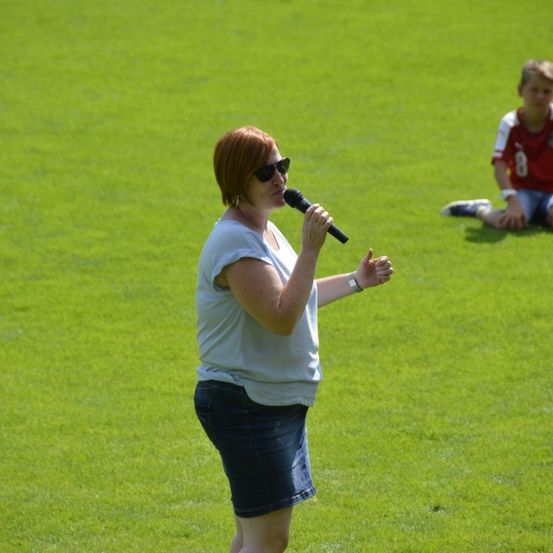 Eine Frau mit roten Haaren steht auf einem Rasenfeld und hält ein Mikrofon. Sie scheint zu singen oder zu sprechen. Ein Junge in einem roten Trikot mit der Nummer 8 sitzt auf dem Gras hinter ihr.