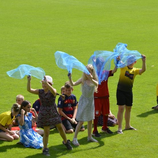 Kinder versammeln sich auf einem Feld, einige stehen und halten blaue Netze, während andere sitzen. Sie sind in verschiedenen Farben gekleidet und scheinen an einer Aktivität beteiligt zu sein.