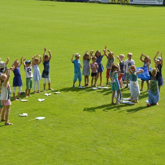 Eine Gruppe von Kindern steht im Kreis auf einem grünen Feld. Sie halten Papierteller und scheinen zu tanzen.