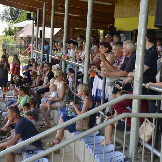 Eine Menschenmenge sitzt auf den Tribünen eines Stadions, einige machen Fotos. Einige stehen in der Nähe der Geländer, während andere nach unten schauen. Einige von ihnen halten Babys. An Stangen sind Lautsprecher montiert.
