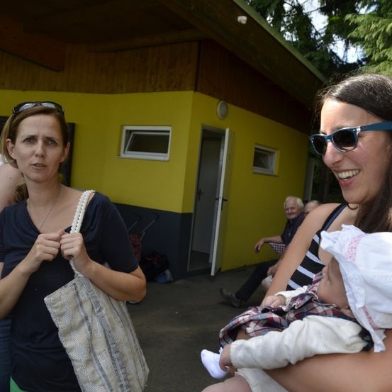 Zwei Frauen und ein Baby stehen vor einem gelben Gebäude mit offener Tür, ein Mann sitzt in der Nähe.