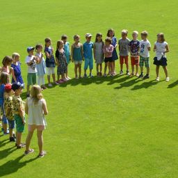 Eine Gruppe von Kindern steht im Kreis auf einem grünen Feld, wahrscheinlich in einer Schulaktivität. Sie tragen verschiedene Sommerkleidung.