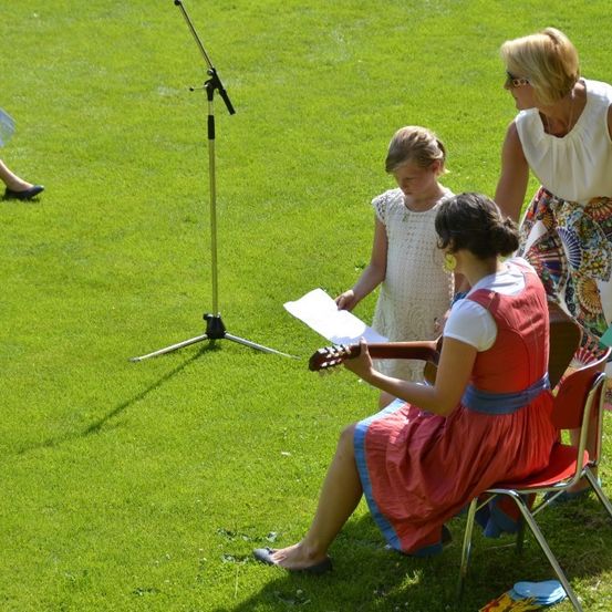 Drei Frauen und ein junges Mädchen sind auf einem Rasenfeld. Eine Frau spielt Gitarre, während das Mädchen ein Papier in der Hand hält. Eine weitere Frau steht hinter ihnen mit einem Papier in der Hand. Ein Mikrofonstativ ist in der Nähe.