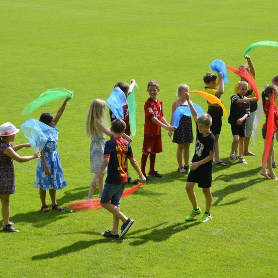 Eine Gruppe von Kindern und Jugendlichen spielt auf einem grünen Feld mit bunten Bändern, einige tragen Turnschuhe und Hüte.