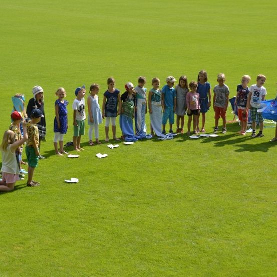 Eine Gruppe von Kindern steht in einem Kreis auf einem grünen Feld und hält Pappteller. Einige tragen Hüte und Brillen. Ein Kind kniet auf dem Boden.