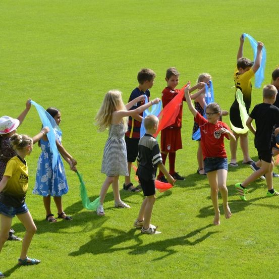 Eine Gruppe von Kindern spielt auf einem Grasfeld und hält bunte Bänder. Sie tragen Freizeitkleidung und einige haben eine Brille. Sie scheinen einen koordinierten Tanz zu machen.